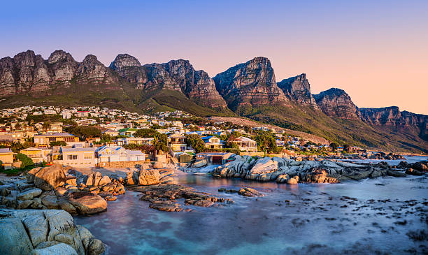 A long exposure at sunset captures the crystal clear turquoise water turning silky smooth at Beta Beach, Bakoven, with the Twelve Apostles mountain range as a backdrop. Cape Town, South Africa